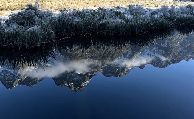 雪山与云朵倒映在平静水面，岸边是覆霜草木